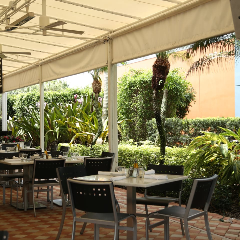 Outdoor patio dining area with tables, chairs, and lush tropical plants under a white awning.