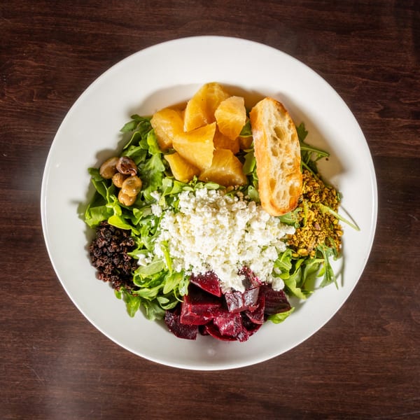 Overhead view of a vibrant salad with arugula, oranges, feta, beets, olives, and a baguette slice.