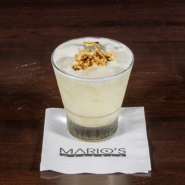 A creamy, light-colored cocktail with toasted coconut flakes on top, resting on a Mario's Osteria napkin.