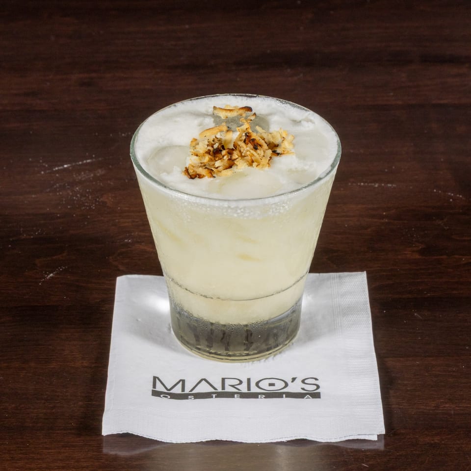 A creamy, light-colored cocktail with toasted coconut flakes on top, resting on a Mario's Osteria napkin.