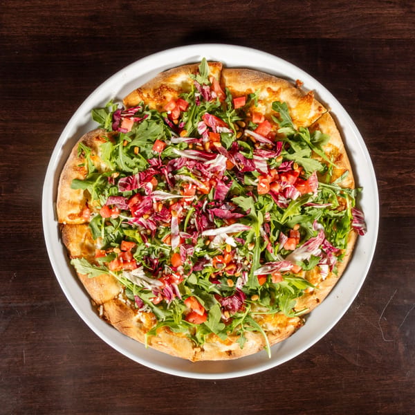 Overhead view of a pizza topped with fresh arugula, radicchio, diced tomatoes, and pine nuts.