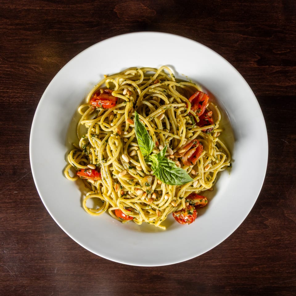 Spaghetti with pesto, cherry tomatoes, pine nuts, and fresh basil on a white plate.