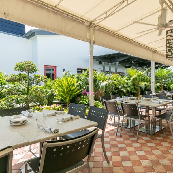 Outdoor patio dining area with set tables, black chairs, and lush green plants under a white awning.