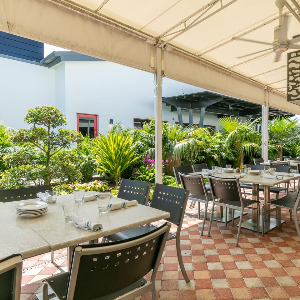 Outdoor patio dining area with set tables, black chairs, and lush green plants under a white awning.