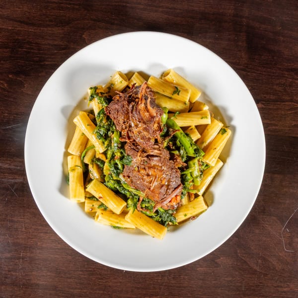 Rigatoni pasta with braised short rib and broccoli rabe in a white bowl on a wooden table.