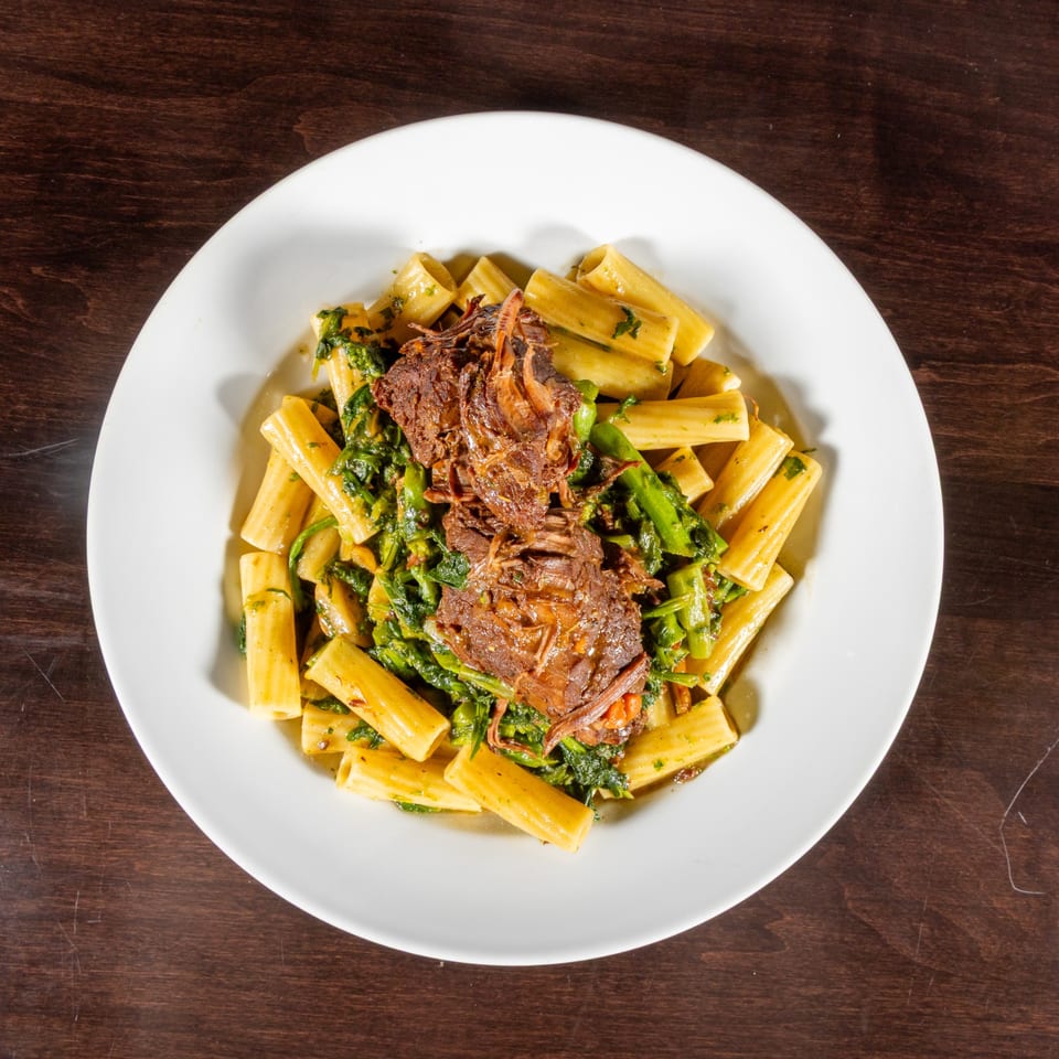 Rigatoni pasta with braised short rib and broccoli rabe in a white bowl on a wooden table.