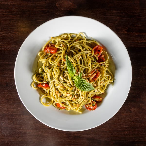 Spaghetti with pesto, cherry tomatoes, pine nuts, and a basil leaf on a white plate.