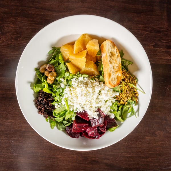 A vibrant salad with arugula, beets, feta, oranges, olives, pistachios, and toasted bread on a white plate.