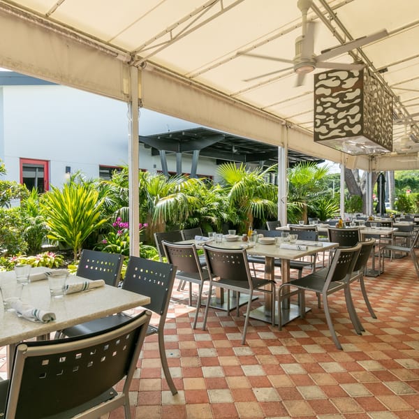 Outdoor restaurant patio with set tables, black chairs, a checkered floor, and lush green plants.