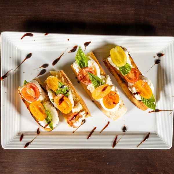 Four bruschetta pieces with cheese, red and yellow tomatoes, basil, and balsamic glaze on a white plate.