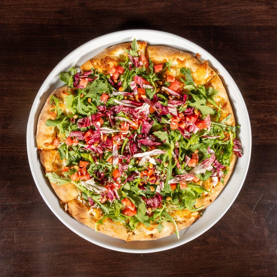 Overhead view of a pizza topped with fresh arugula, radicchio, diced tomatoes, and pine nuts.